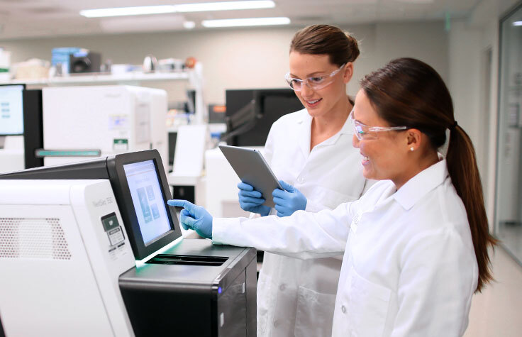 Two women, wearing lab coats and safety gear, operating sequencing equipment in a lab.