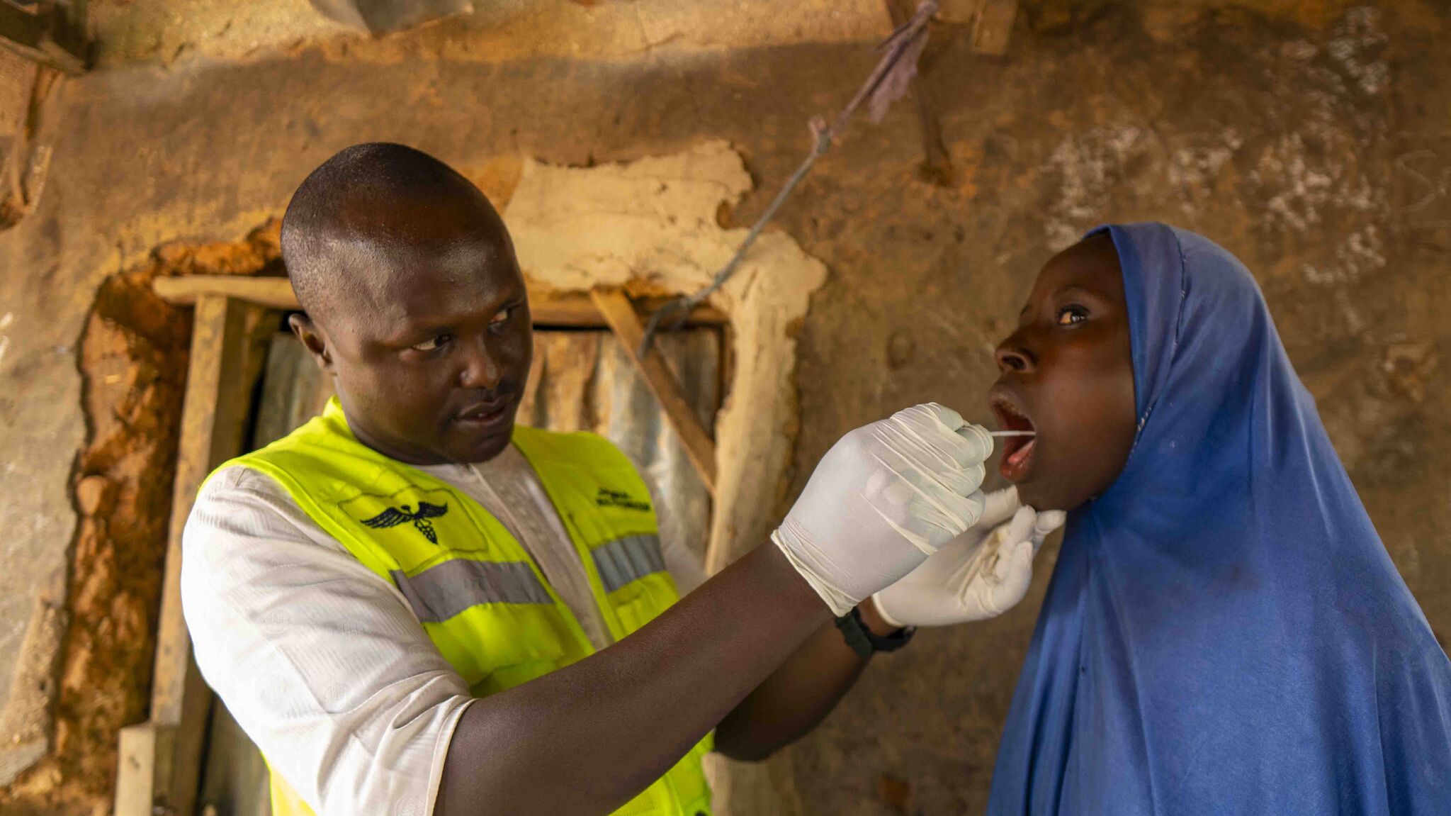 A young African doctor wearing a white coat, gloves, and stethoscope writes on a clipboard while a seated male patient rests his arm on a table during an outdoor medical checkup.